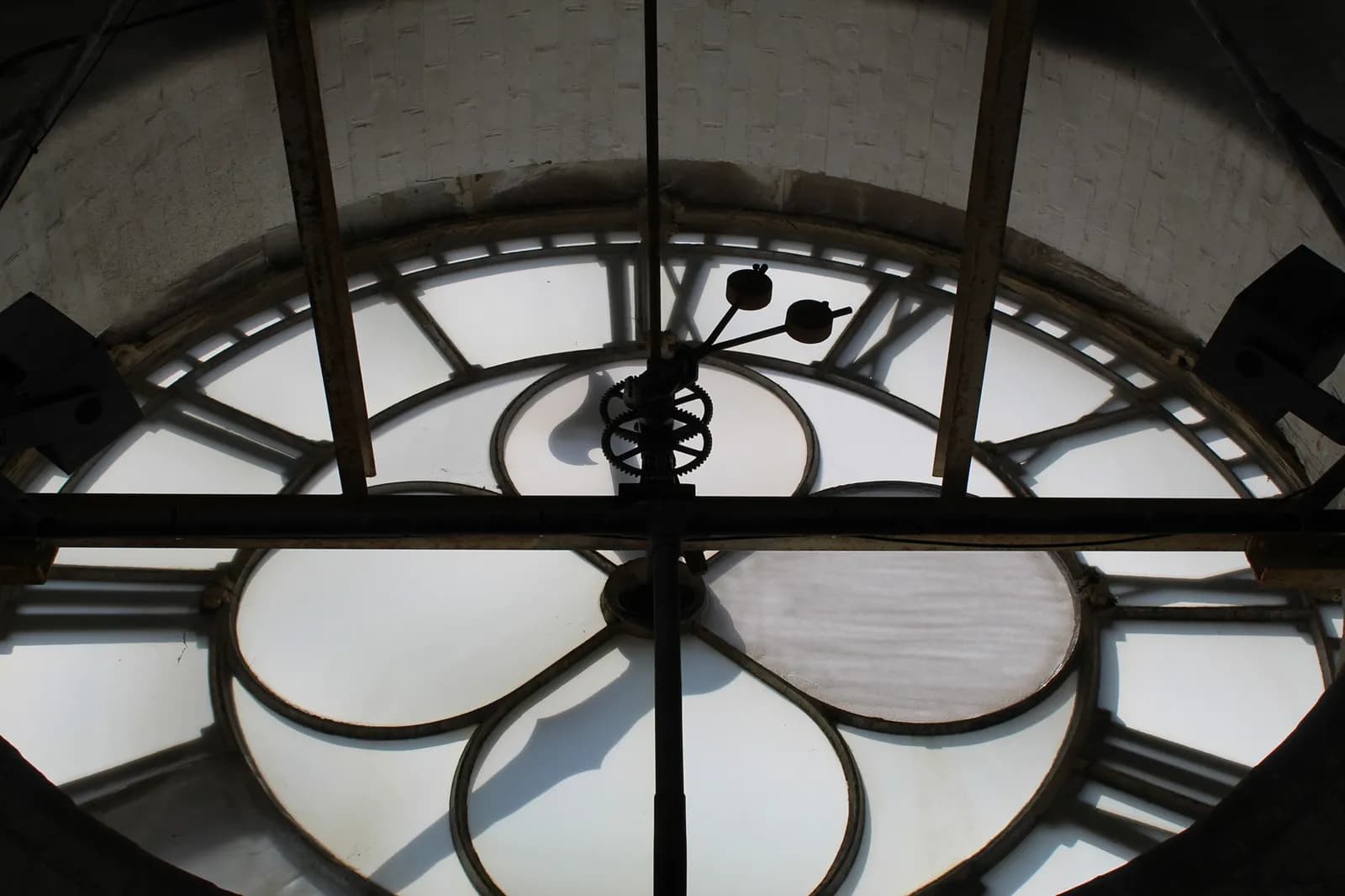 A close-up view from inside a large clock tower, showing the intricate mechanism of gears and hands against the translucent clock face, revealing Roman numerals and structural beams.