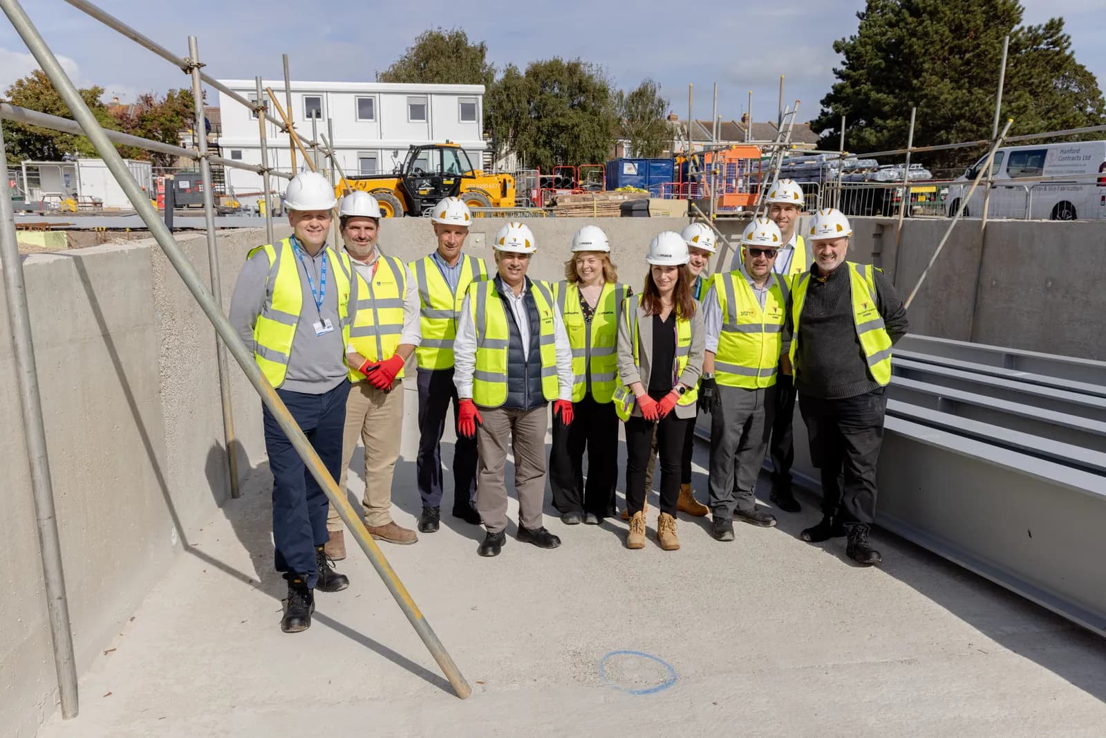 A group of construction workers wearing hard hats and safety vests stand together on a concrete surface at a construction site, with modular buildings and construction equipment visible in the background.