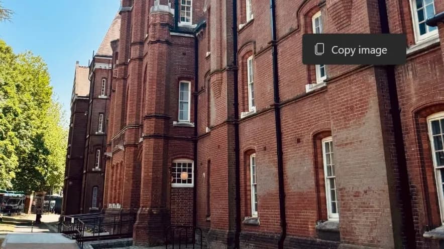 A long, red brick building with many windows extends diagonally from the left to the right of the frame, with green trees visible on the left side under a clear blue sky.