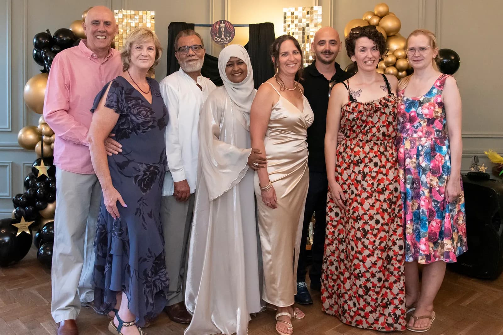 A group of eight adults, four men and four women, stand together smiling in formal attire in front of a decorated wall with golden and black balloons and an Oscars sign.