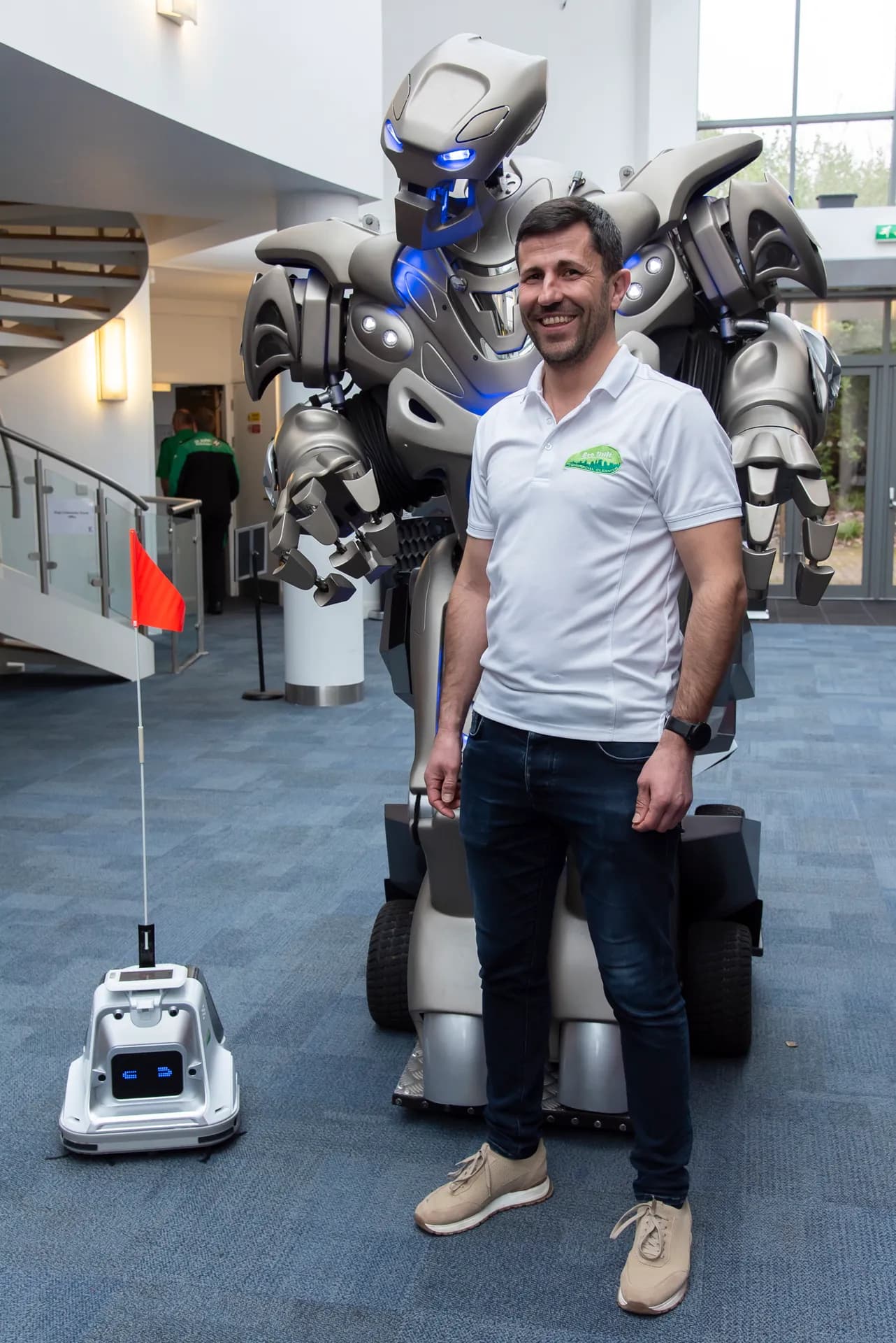 A man in a white polo shirt stands next to a large, futuristic-looking robot, with a smaller robotic device carrying a red flag to their left.